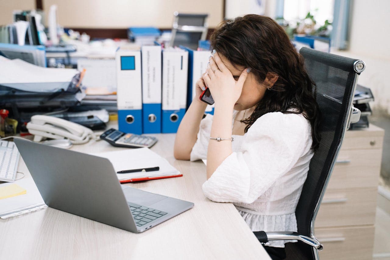 Stressed office worker sitting at a desk with a laptop, holding a phone and covering her face